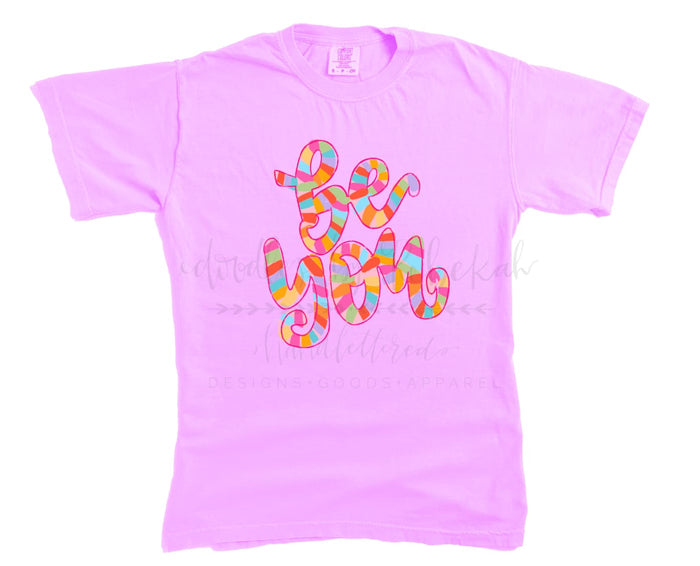 A pink unisex t-shirt with a colorful text design on the front.