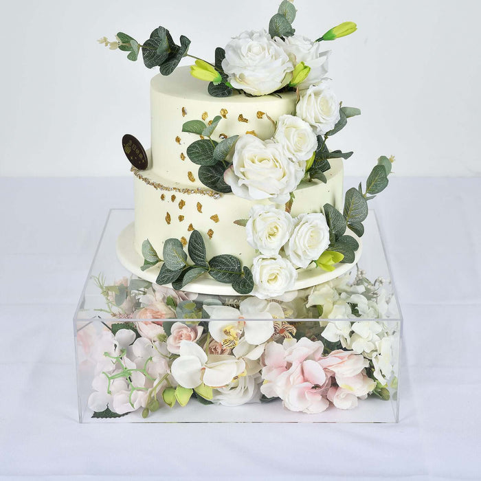 Elegant three-tier cake adorned with white roses and greenery, displayed in a clear box.