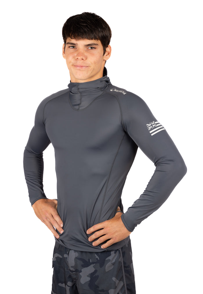 Male model wearing the Waihana Anthracite Carvico Hooded Surf Rashguard in charcoal gray, featuring a built-in face gaiter and Hawaiian flag sleeve logo, paired with camo board shorts.