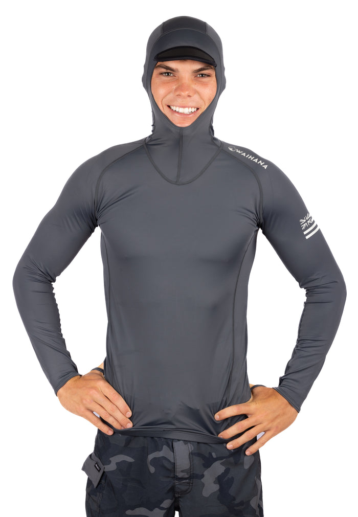 Male model wearing a Waihana Anthracite Carvico Hooded Surf Rashguard in grey with integrated hood and brim, Hawaiian flag sleeve logo, paired with camo shorts on a white studio background.