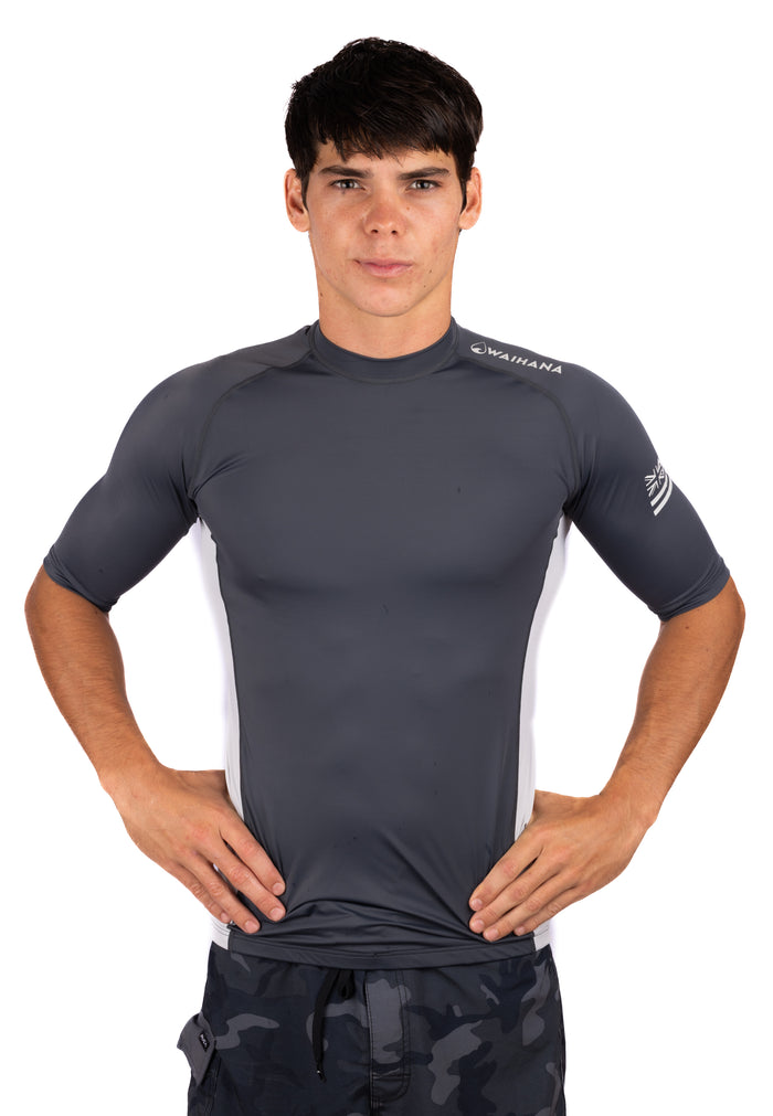 Male model wearing a Waihana Anthracite short sleeve rashguard with white side panels and Waihana logo on the chest, paired with camo board shorts, on a white studio background.
