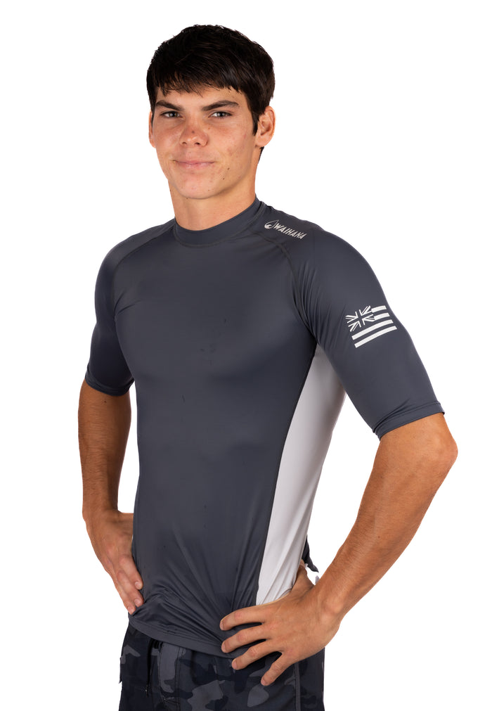 Male model wearing the Waihana Anthracite Short Sleeve Rashguard in dark charcoal grey with a light grey side panel and Hawaiian flag sleeve graphic, shown from a slight angle against a white background.