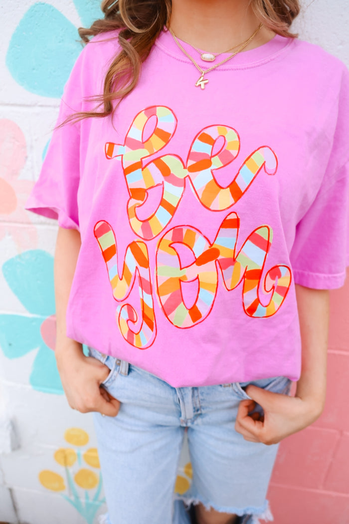 A person wearing a pink t-shirt with the text 'BE YOU' in a hand-drawn style font in multicolor.
