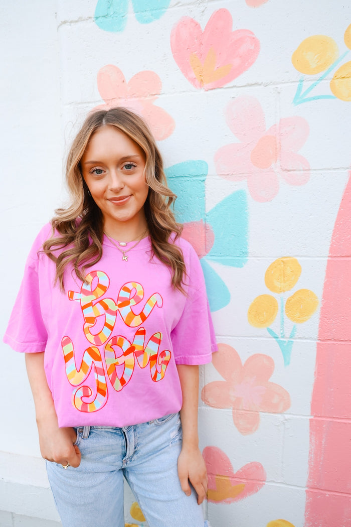 Girl wearing a pink shirt with colorful hand drawn style graphic 'be you' against a flower mural
