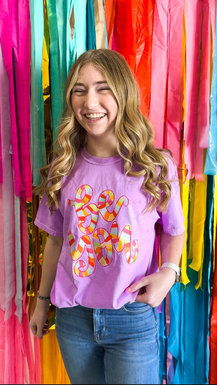 A person smiling and wearing a purple t-shirt with hand-drawn lettering and designs.