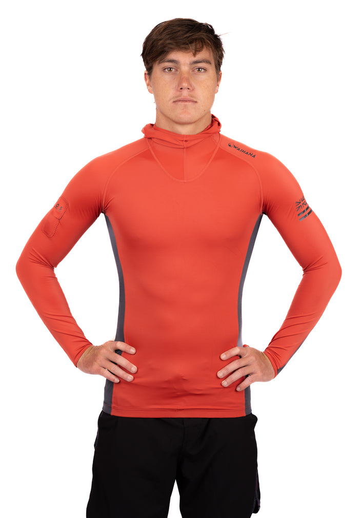 Male model wearing the Waihana Canyon Hooded Voyager Rashguard in orange-red with grey side panels and long sleeves, standing hands on hips against a white studio background.