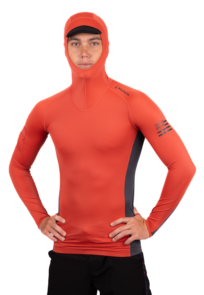 Male model wearing the Waihana Canyon Hooded Voyager Rashguard in orange-red with grey side panels and an integrated hood with black brim, hands on hips against a white background.