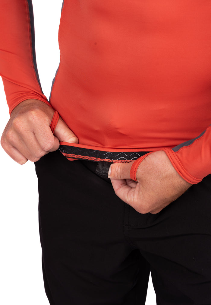 Close-up of a man stretching the hem of the Canyon Hooded Voyager Rashguard in red-orange, revealing the textured elastic waistband attachment detail against black wetsuit pants.