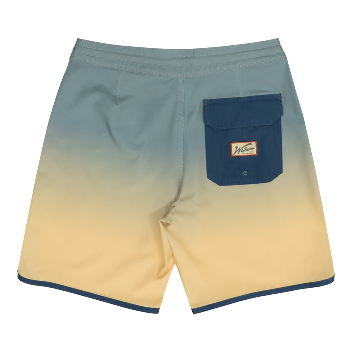 Back view of Waihana Dawn Patrol W1 board shorts with a blue-to-yellow ombre gradient, navy flap back pocket with branded logo patch, and navy trim along the hem.