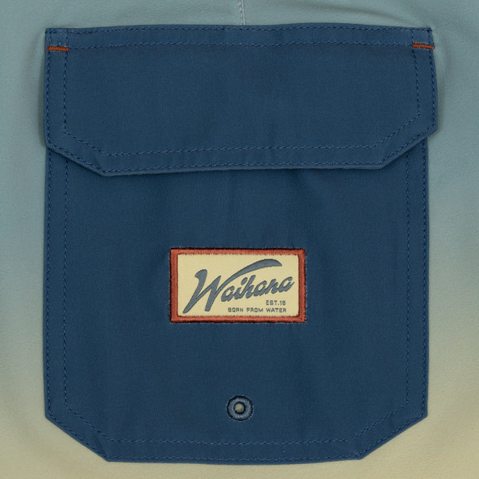 Close-up of the dark blue cargo pocket on the Waihana Dawn Patrol W1 Board Shorts, showing the embroidered cream and red Waihana brand patch and a drain grommet at the bottom.