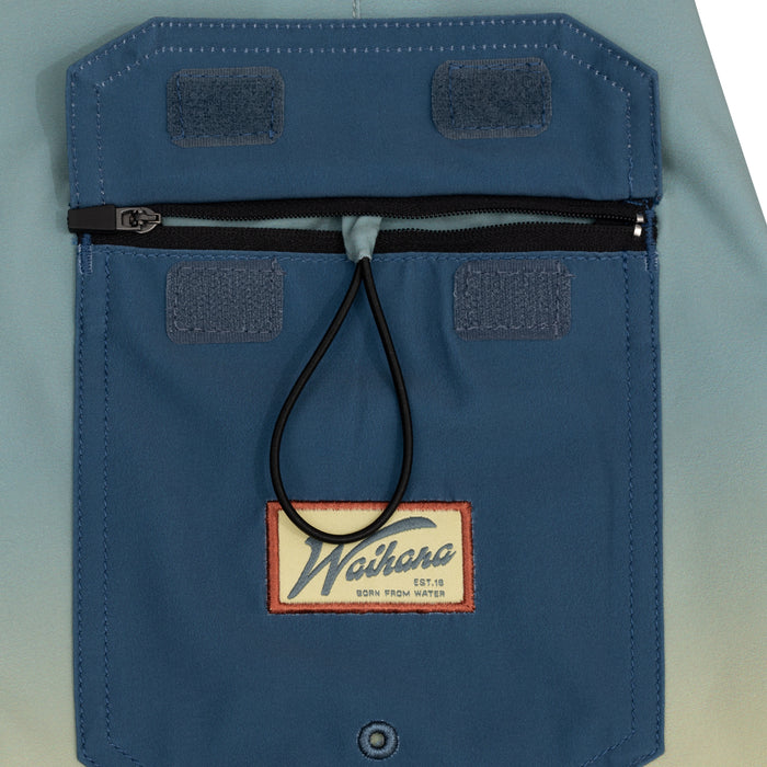 Close-up of the rear zippered pocket on the Waihana Dawn Patrol W1 Board Shorts in blue ombre, featuring Velcro panels, a rubber cord loop, and an embroidered Waihana 'Born From Water' patch.