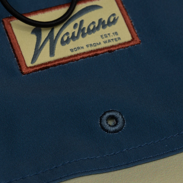Close-up of a navy blue Waihana Dawn Patrol W1 board short showing the embroidered 'Born from Water' patch label and a stitched buttonhole eyelet detail.