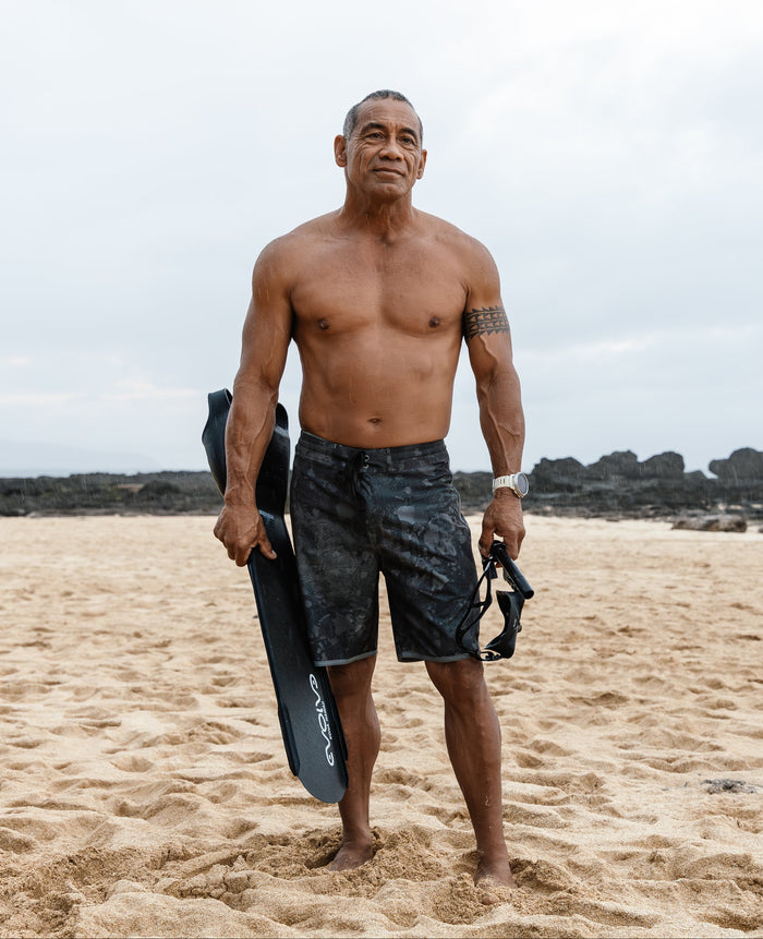 Shirtless male spearfisher stands confidently on a sandy beach holding Evolve freediving fins and a dive mask, wearing Waihana Island Aloha W1 camo board shorts with rocky coastline in the background.
