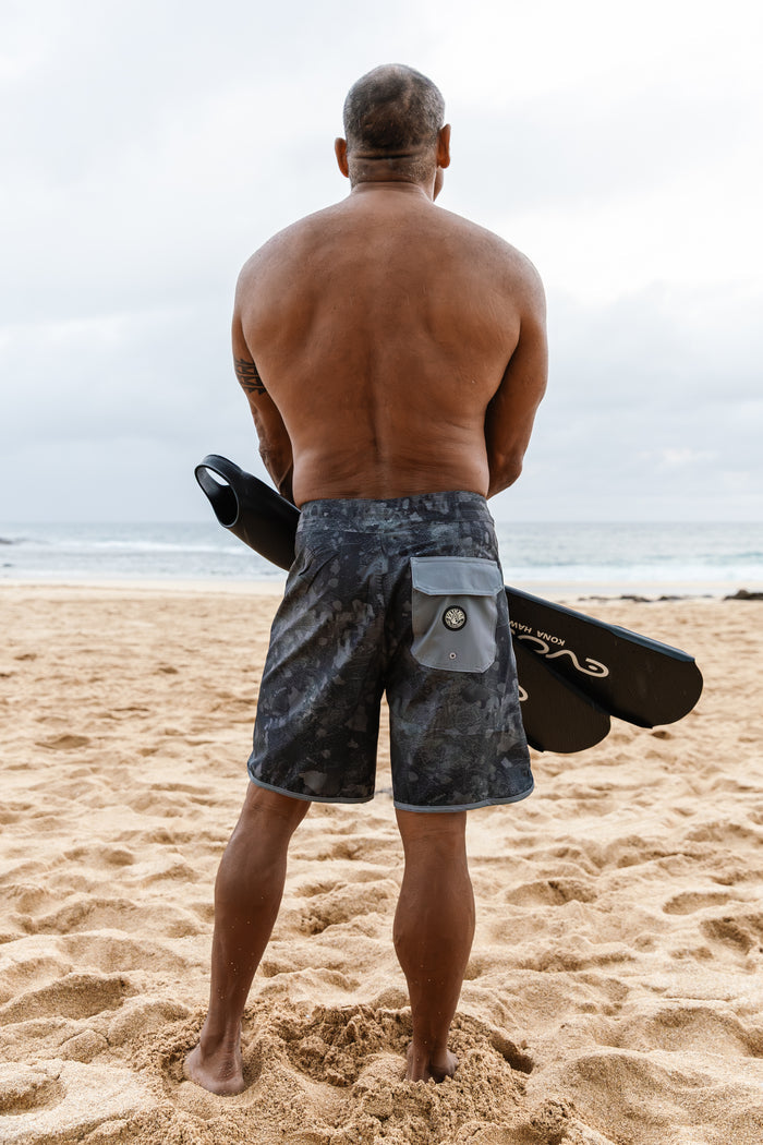 Man standing on a sandy beach from behind wearing Waihana Island Aloha W1 camo board shorts and holding black freediving fins, with the ocean visible in the background.