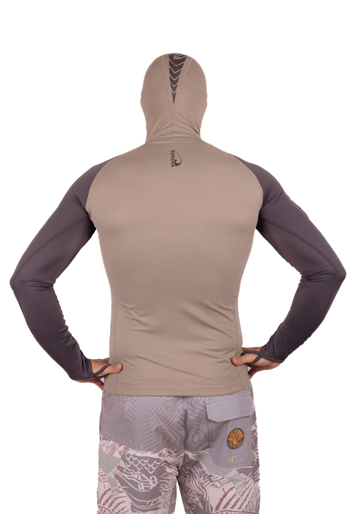 Back view of a male model wearing the Waihana Mana Hooded Spearfishing Rashguard in tan with dark grey contrast sleeves and a fitted hood featuring chevron graphic detailing, paired with Waihana patterned board shorts.