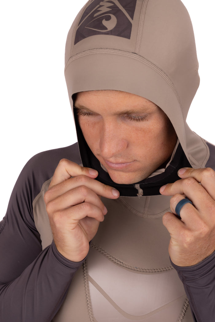 Male model adjusting the fitted hood of the Waihana Mana Hooded Spearfishing Rashguard in tan and grey, shown in close-up studio detail.