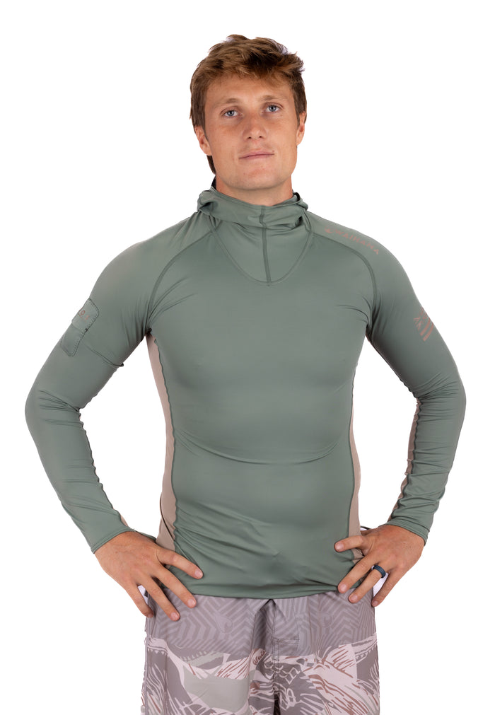 Male model wearing the Waihana Olive Hooded Voyager Rashguard, a long-sleeve sage green rashguard with tan side panels and a half-zip hood, paired with grey patterned board shorts.