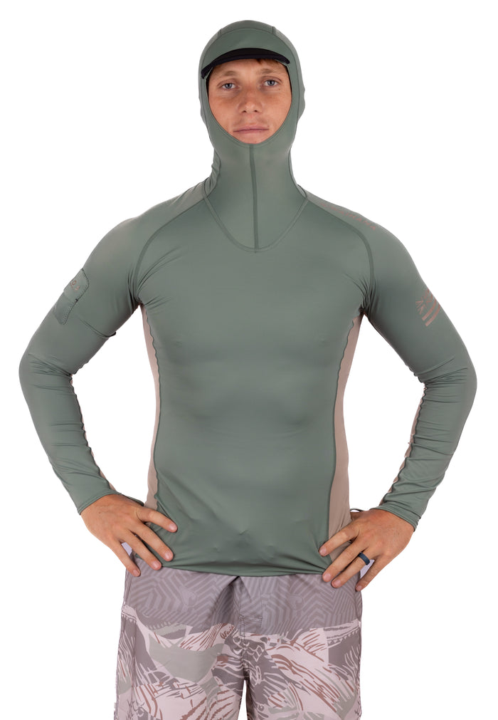 Male model wearing the Waihana Olive Hooded Voyager Rashguard, a fitted long-sleeve sun protection top with hood and contrasting side panels, paired with patterned board shorts.