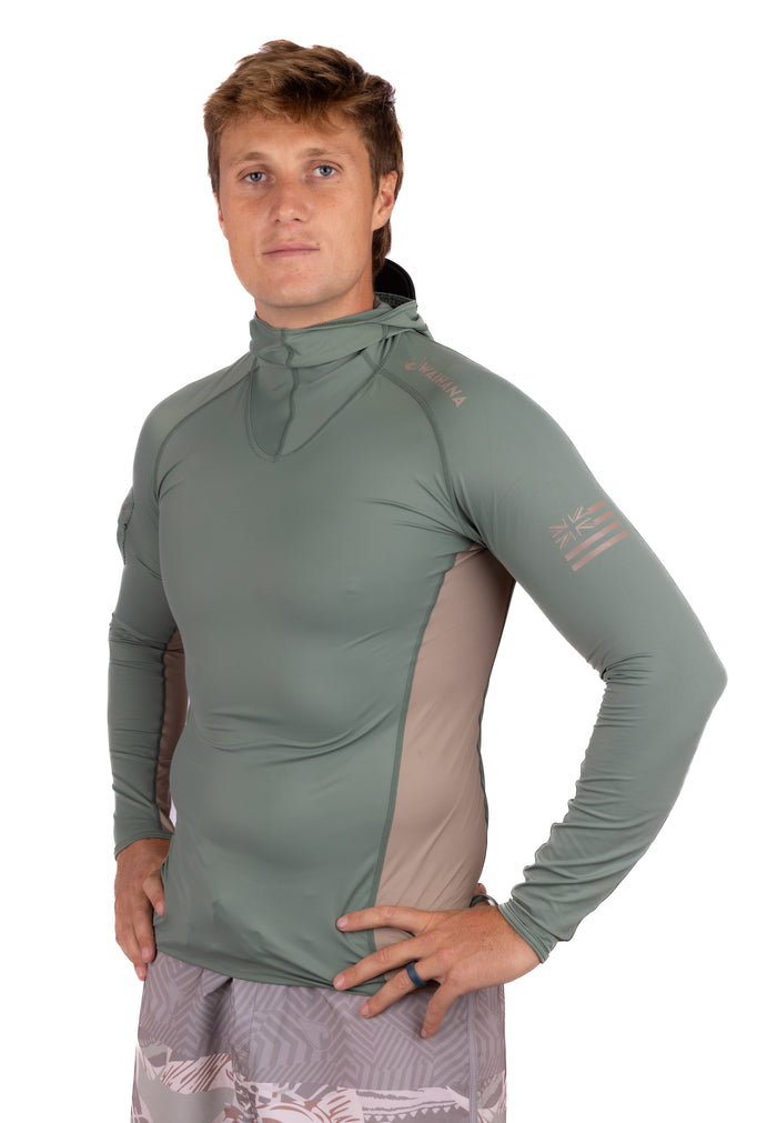 Male model wearing the Waihana Olive Hooded Voyager Rashguard in sage green with tan side panels, built-in neck gaiter, and Hawaiian flag sleeve graphic, paired with printed board shorts.