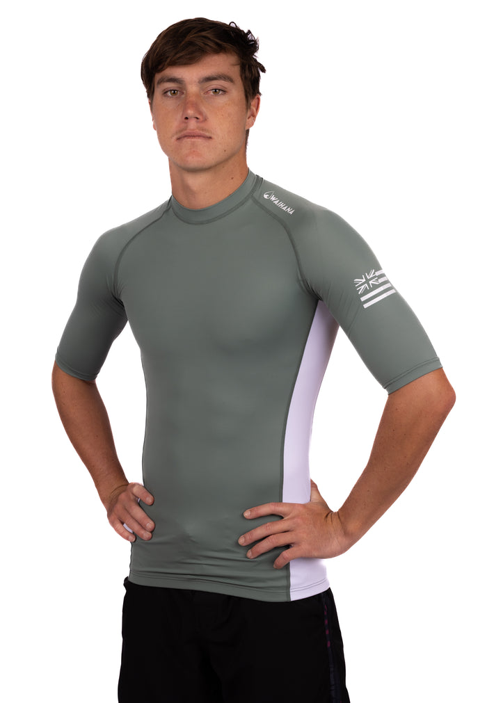 Male model wearing the WAIHANA Olive Short Sleeve Rashguard with white side panels and Hawaiian flag sleeve graphic, shown from a slight angle against a white studio background.