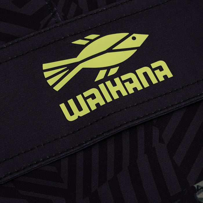 Close-up of the lime green Waihana fish logo and wordmark printed on the waistband of black Tropicam W1 board shorts with a subtle tribal pattern.