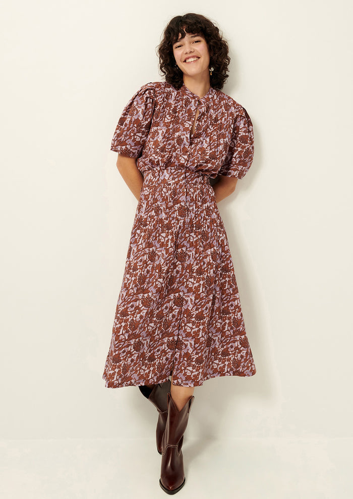 A woman standing wearing an abstract printed purple and rust colored button up top with short sleeves, matching skirt, and brown boots..